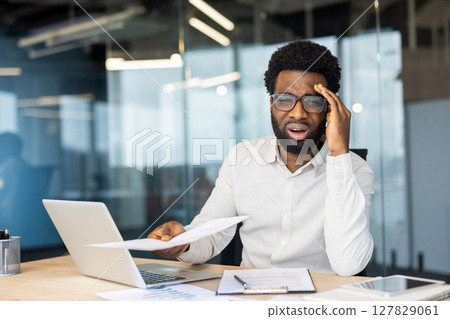 An African-American businessman with a headache, overwhelmed by paperwork in an office setting, stressed and frustrated. An African-American businessman with a headache, overwhelmed by paperwork in an office setting, stressed and frustrated. 127829061