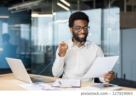An african american man sits at his desk, ecstatic, holding papers, laptop, and a tablet are on the table. He appears joyful and has an expression of achievement. An african american man sits at his desk, ecstatic, holding papers, laptop, and a tablet are on the table. He appears joyful and has an expression of achievement. 127829063