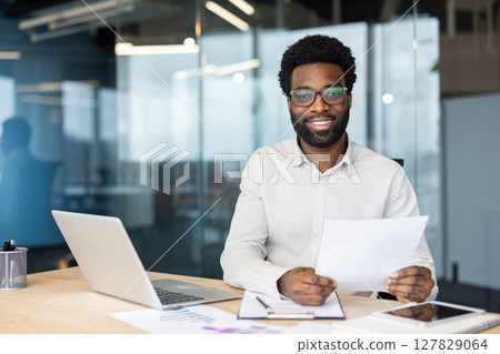 Smiling professional with glasses holds documents while working at his office desk. He looks at the camera with confidence and ease. Smiling professional with glasses holds documents while working at his office desk. He looks at the camera with confidence and ease. 127829064