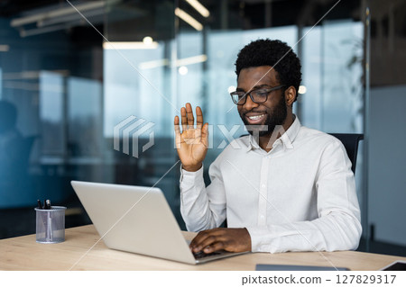 A smiling African American man in glasses waves during a video call on his laptop in a modern office, connecting with colleagues or clients. A smiling African American man in glasses waves during a video call on his laptop in a modern office, connecting with colleagues or clients. 127829317