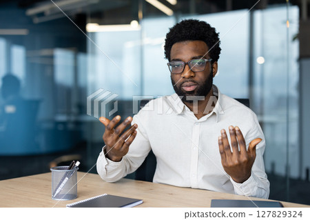 An African American man wearing glasses seriously communicates during a video call from his modern office, gesturing with his hands An African American man wearing glasses seriously communicates during a video call from his modern office, gesturing with his hands 127829324