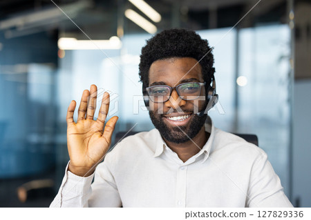 A cheerful man with glasses and a headset waves, expressing a friendly welcome or greeting. He smiles in an office setting. 127829336