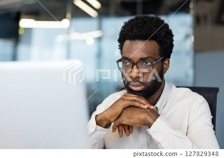 A focused, man wearing glasses looks intently at a computer screen, deep in thought at work. A focused, man wearing glasses looks intently at a computer screen, deep in thought at work. 127829348
