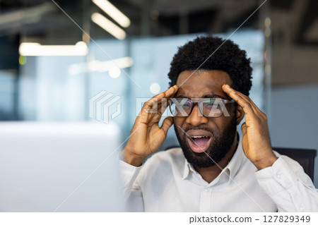 A Black man in glasses reacts to bad news on his computer, with an expression of frustration and hands to his temples 127829349