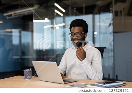 A smiling Black businessman in glasses sits at a desk with a laptop, ready for work. Modern office with glass walls is visible in the background. 127829361