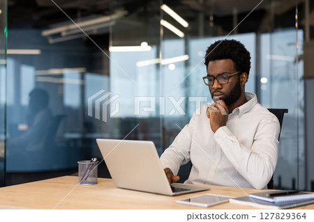 African American businessman thinking laptop work. Capturing thoughtful expression, business attire, office ambiance. Represents focus, technology usage, corporate environment, decision making. 127829364