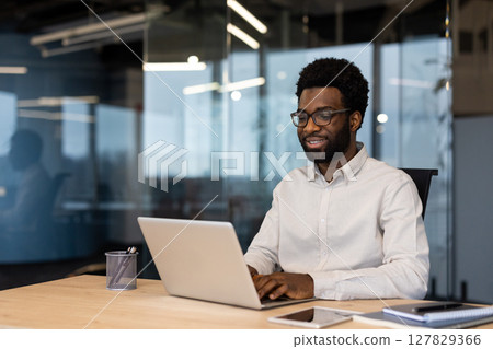 Young successful businessman working on laptop inside office, man in light shirt smiling contentedly, typing on laptop keyboard at workplace. Young successful businessman working on laptop inside office, man in light shirt smiling contentedly, typing on laptop keyboard at workplace. 127829366