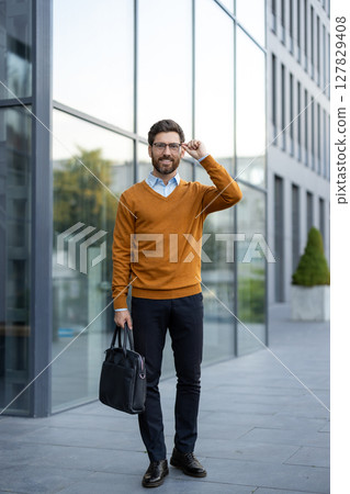 A smiling businessman with glasses adjusts his frames while holding a briefcase outside a modern office building. He wears a smart casual attire. A smiling businessman with glasses adjusts his frames while holding a briefcase outside a modern office building. He wears a smart casual attire. 127829408