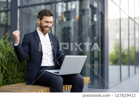 A successful businessman celebrates his achievement while working on his laptop outdoors, exuding joy and satisfaction. 127829412
