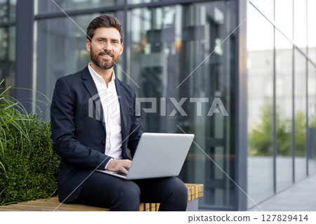 A smiling businessman sits outside, working on a laptop. He wears a suit and is positioned in front of a modern building, suggesting a professional setting. 127829414