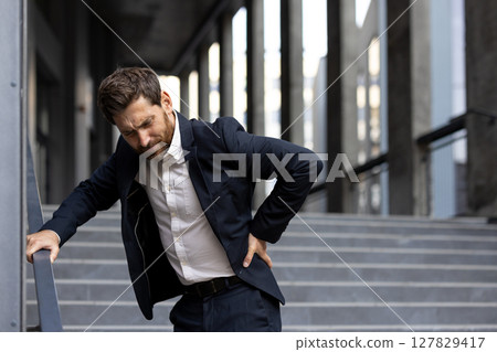 A businessman grimaces in pain as he clutches his back while standing on stairs, highlighting a potential work injury or health concern. 127829417