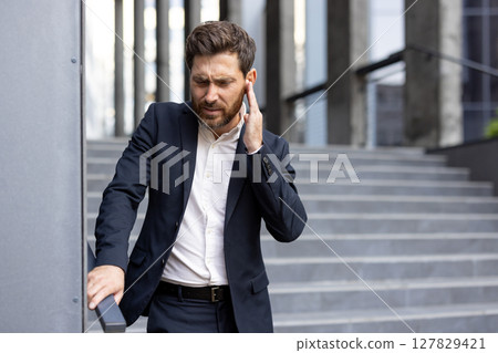 A concerned businessman in a dark suit touches his ear, standing near a modern building's staircase, showing visible stress. A concerned businessman in a dark suit touches his ear, standing near a modern building's staircase, showing visible stress. 127829421