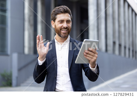 A smiling businessman holds a tablet outdoors, waving during a video call. The urban background is blurred, but the focus is clear. 127829431