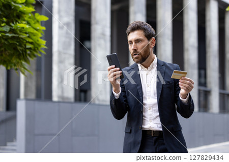 A businessman looks shocked while holding a credit card and his phone, possibly due to a financial issue. A businessman looks shocked while holding a credit card and his phone, possibly due to a financial issue. 127829434