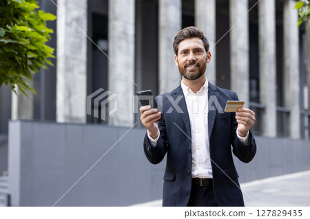 A smartly dressed businessman smiling while holding a credit card and his phone, outdoors near a modern building. 127829435