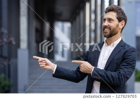 A smiling businessman in a dark suit points both fingers to the right, near an office building, ready to lead. A smiling businessman in a dark suit points both fingers to the right, near an office building, ready to lead. 127829703
