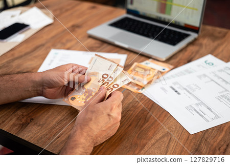 Man counting euro banknotes while managing household expenses and utility bills. Focus on financial planning, tax calculations, and online payment management Man counting euro banknotes while managing household expenses and utility bills. Focus on financial planning, tax calculations, and online payment management 127829716