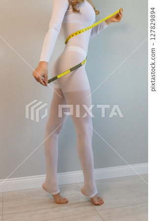 woman in a white suit for LPG massage holds a centimeter in her hands. Anti-cellulite body care concept. Gray background woman in a white suit for LPG massage holds a centimeter in her hands. Anti-cellulite body care concept. Gray background 127829894
