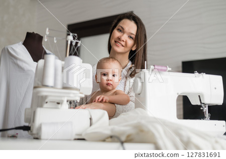 A young woman sews in the kitchen and holds a small child. Mom teaches her little son to sew on a sewing machine. Self-isolation homework 127831691