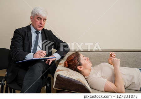 Caucasian woman lies on a couch at a reception with a psychotherapist. An elderly man works as a psychiatrist Caucasian woman lies on a couch at a reception with a psychotherapist. An elderly man works as a psychiatrist 127831875