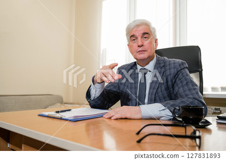 Confident elderly gray-haired man in a plaid suit sits at his desk in the office Confident elderly gray-haired man in a plaid suit sits at his desk in the office 127831893