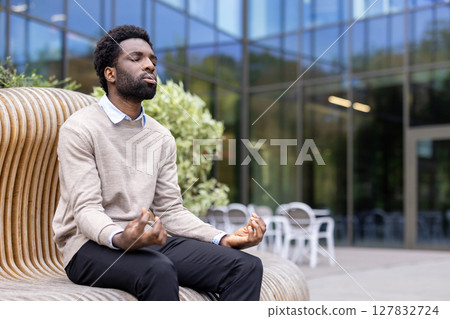 An african american man meditates peacefully outdoors, finding calm in the natural light near a modern building. 127832724