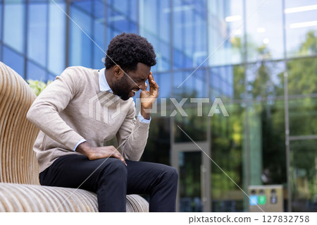 A man sits outdoors, expressing a headache, stress or pain in front of an office building, grasping his head with his hand. He appears to be in distress. A man sits outdoors, expressing a headache, stress or pain in front of an office building, grasping his head with his hand. He appears to be in distress. 127832758