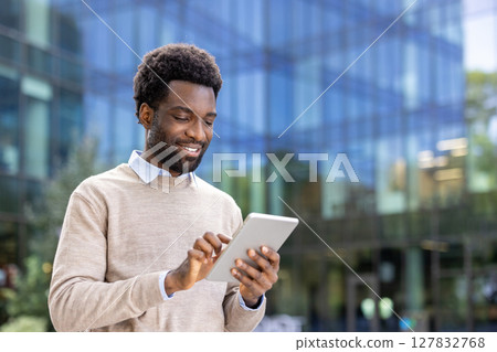 A smiling businessman looks at his tablet while standing outdoors near a modern office building. 127832768