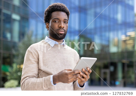 An african american man in a light sweater, holding a tablet, posing in front of an office building, looking at the camera. 127832769