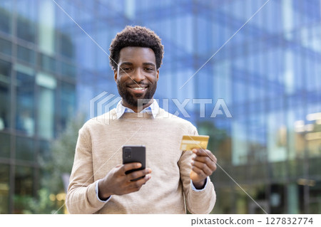 A cheerful man holding a smartphone and credit card, smiling towards the camera in an outdoor setting. He is likely engaged in a transaction. A cheerful man holding a smartphone and credit card, smiling towards the camera in an outdoor setting. He is likely engaged in a transaction. 127832774