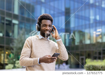 Smiling man wearing headphones holds a phone in front of a modern building with a glass facade. 127832775