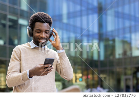 An African-American man smiles while listening to music through headphones, holding his phone outside a modern building. An African-American man smiles while listening to music through headphones, holding his phone outside a modern building. 127832776