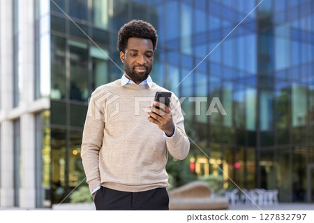 An African American man checks his phone in front of a modern glass building, possibly waiting outside an office. 127832797