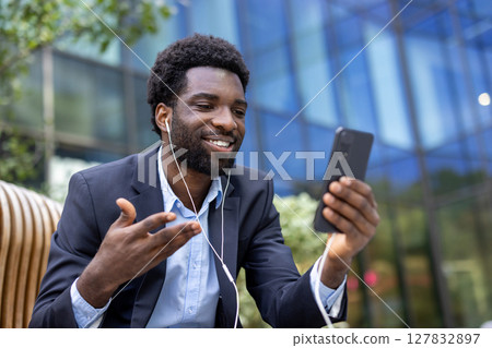 A smiling businessman in a suit is holding a phone and having a video call outdoors. He is using earbuds. A smiling businessman in a suit is holding a phone and having a video call outdoors. He is using earbuds. 127832897