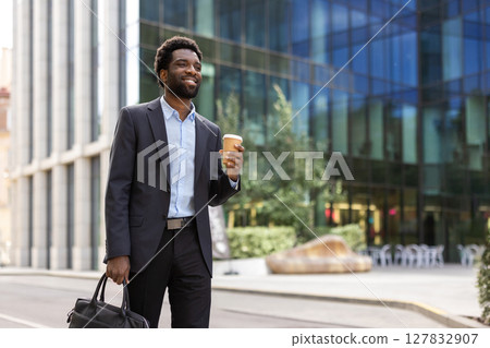 Young successful businessman walking outside office building with cup of hot drink in hand and business bag suitcase. Man in business suit smiling happily. 127832907