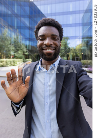 A young, handsome man in a blazer smiles and waves to the camera during a mobile video call in front of a modern office building 127832919