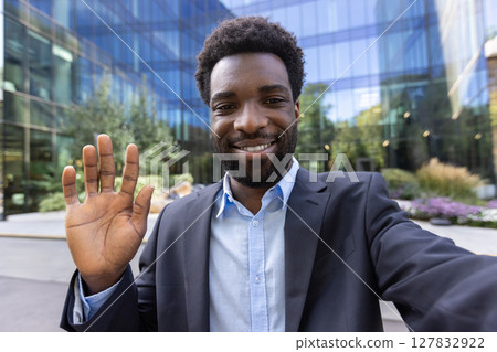 An attractive man in a business suit is waving in front of a modern building. Smiling directly at the camera, outdoors. 127832922