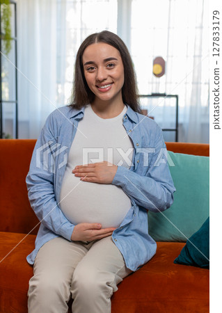 Portrait smiling future mother woman sitting on sofa touching pregnant belly expecting baby at home 127833179