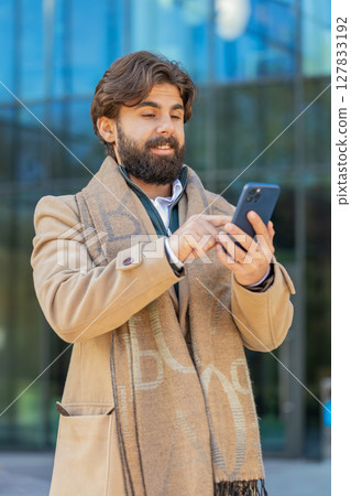 Smiling young man tourist chatting on internet via mobile phone standing on downtown city street Smiling young man tourist chatting on internet via mobile phone standing on downtown city street 127833192