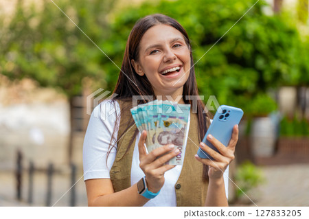 Happy young woman counting money UAH Ukrainian cash, using smartphone calculator app on city street Happy young woman counting money UAH Ukrainian cash, using smartphone calculator app on city street 127833205