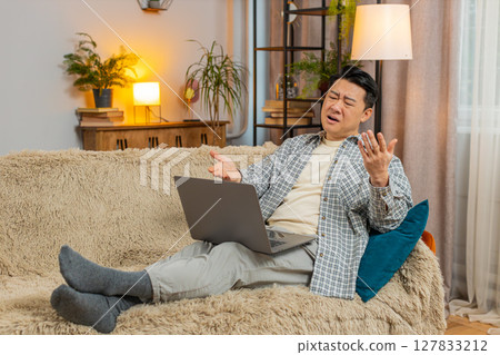Upset displeased Chinese man using laptop computer reading bad news sitting on sofa in living room Upset displeased Chinese man using laptop computer reading bad news sitting on sofa in living room 127833212