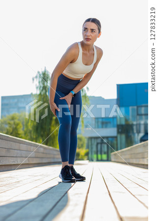Athletic runner woman in sportswear with muscle pain after jogging in urban city park on sunny day 127833219