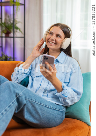 Joyful young woman enjoying listening to music through wireless headphones dancing at home room sofa 127833221