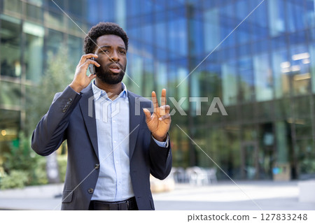 A handsome businessman gesturing while on his phone stands confidently in front of an office building on a bright day. A handsome businessman gesturing while on his phone stands confidently in front of an office building on a bright day. 127833248