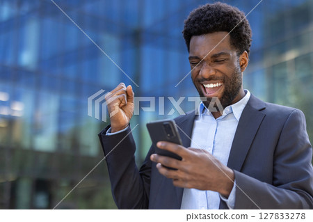 An excited businessman in a suit celebrates with a clenched fist while looking at his phone, with a blurred building background. An excited businessman in a suit celebrates with a clenched fist while looking at his phone, with a blurred building background. 127833278