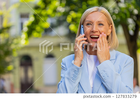 Happy mature Caucasian businesswoman lady smiling laughs while talking on smartphone on city street 127833279
