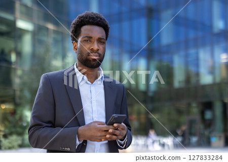 A smartly dressed African-American man holds his smartphone, posing confidently in front of a modern office building. A smartly dressed African-American man holds his smartphone, posing confidently in front of a modern office building. 127833284