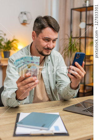 Smiling Caucasian young man counting money cash using smartphone at home office table in living room 127833301