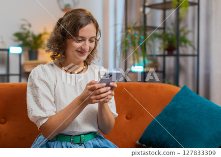 Happy smiling woman sitting on couch using smartphone texting smiling at home living room apartment Happy smiling woman sitting on couch using smartphone texting smiling at home living room apartment 127833309