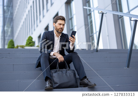 A concerned businessman wearing a suit checks his phone while sitting on outdoor steps near a modern building. He holds a briefcase. 127833313